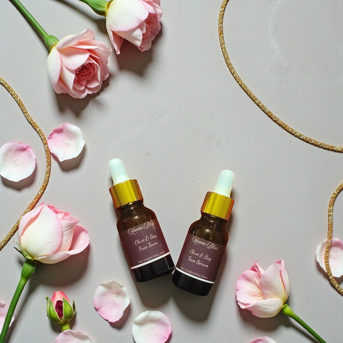 Haven Bliss Skincare Clove & Bay Ritual Duo, two amber glass dropper bottles with gold accents, surrounded by pink roses and petals.